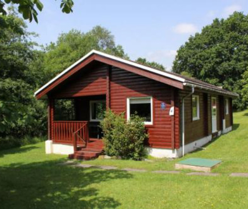 wooden cabin surrounded by trees with a porch and green lawn perfect for three guests