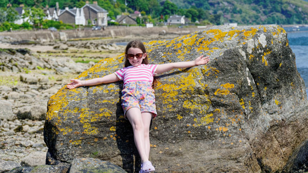 young girl in sunglasses relaxing on a large rock by the seaside with colorful lichen enjoying a sunny day near the beach and coastal homes 9 adventures in nature 9 summer fun